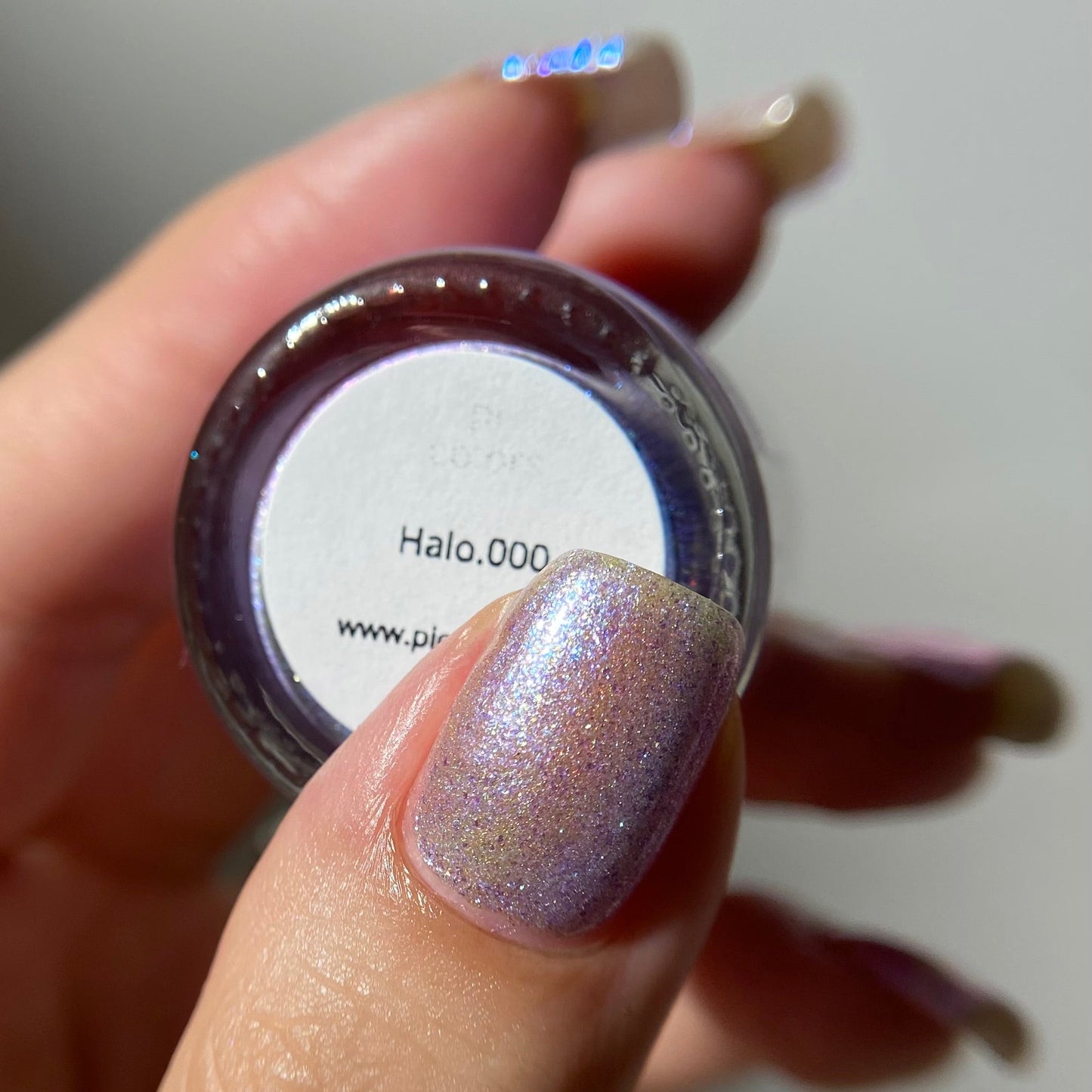A close-up of a hand holding a small round pot of nail polish or pigment. The product color is a shimmering holographic lavender with iridescent sparkle, and the label reads 'Halo.