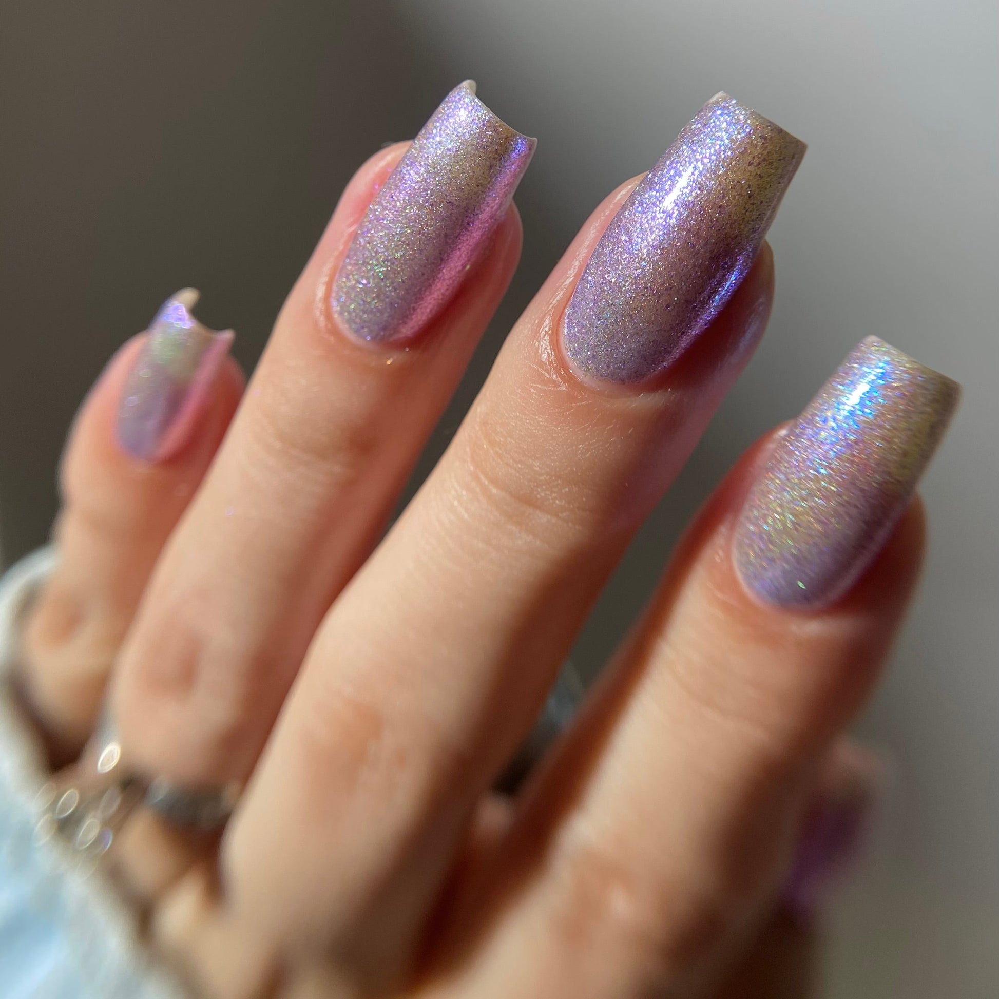 A close-up of a hand with long, square-shaped nails painted in a shimmering iridescent lavender and gold holographic nail polish.