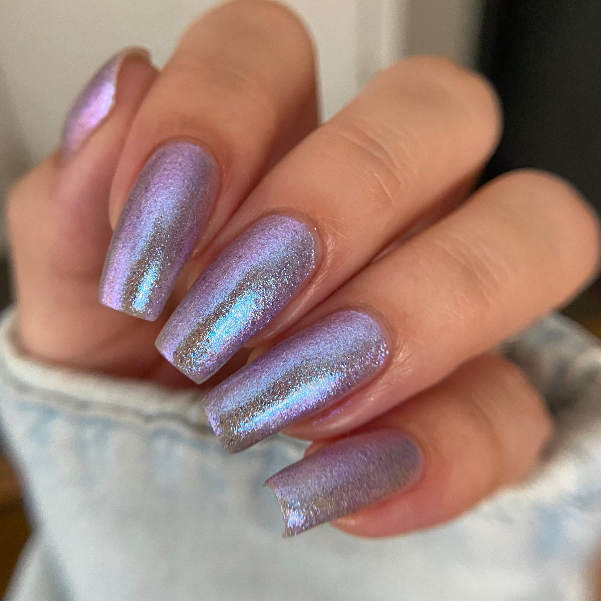 A close-up of a hand with manicured nails painted in a shimmering iridescent lavender-purple polish with gold glitter, wearing a light denim jacket.