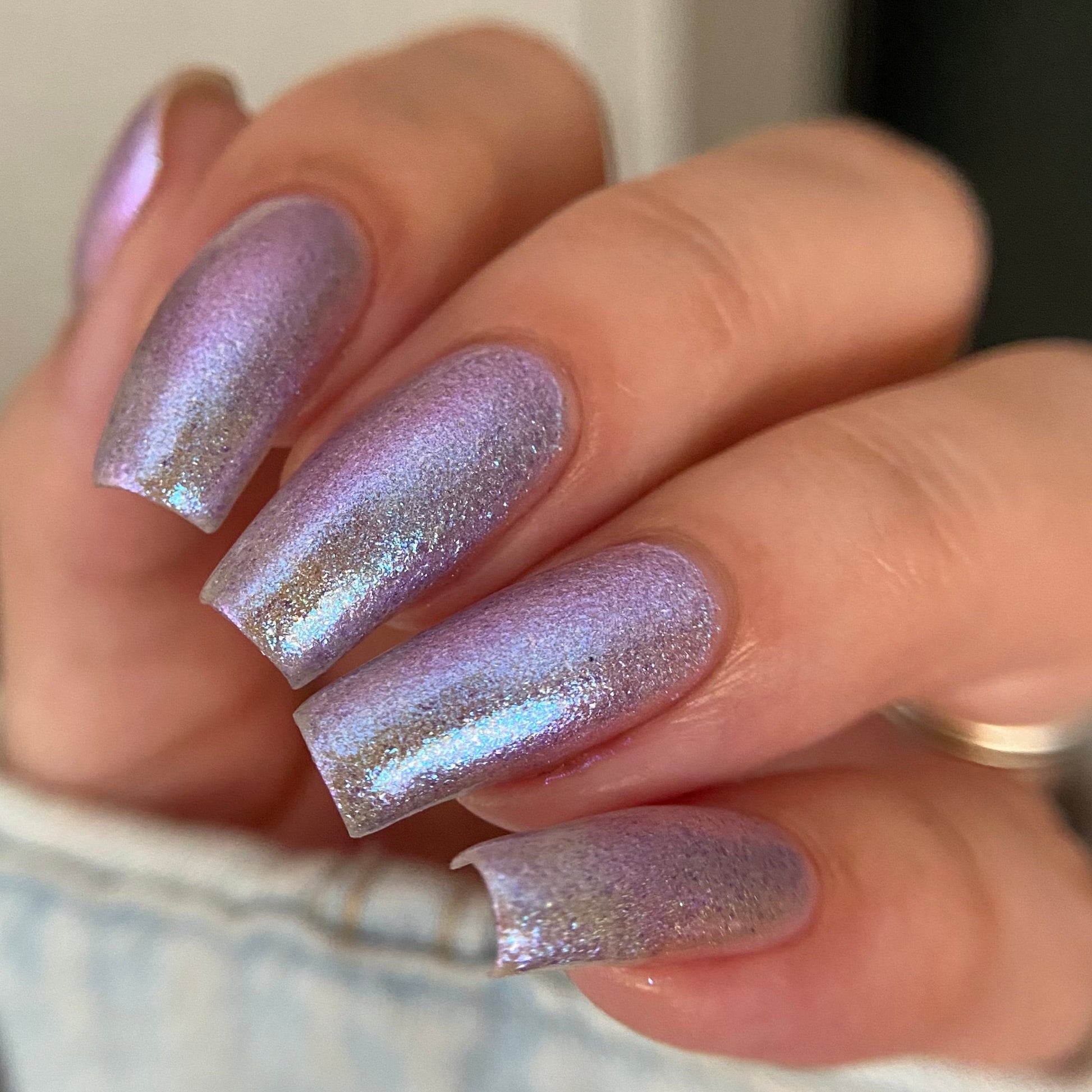 A close-up of a hand with manicured nails painted in a shimmering iridescent lavender-purple polish with fine gold glitter, wearing a light denim sleeve.