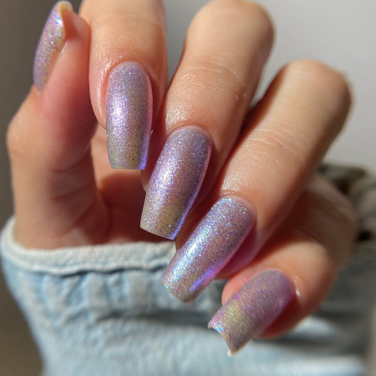 A close-up of a hand with long, almond-shaped nails painted in a shimmering iridescent lavender and silver holographic nail polish, wearing a light blue denim sleeve.