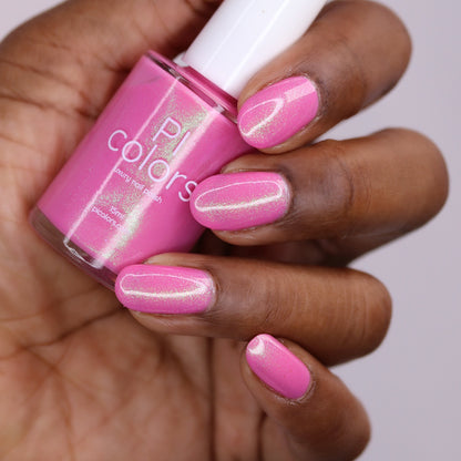 A hand with medium-brown skin holds a bottle of PI Colors' Mint and Summer.100, a handmade fluorescent pink nail polish. The nails are painted pink with a color-shifting shimmer that matches the vibrant polish in the bottle.