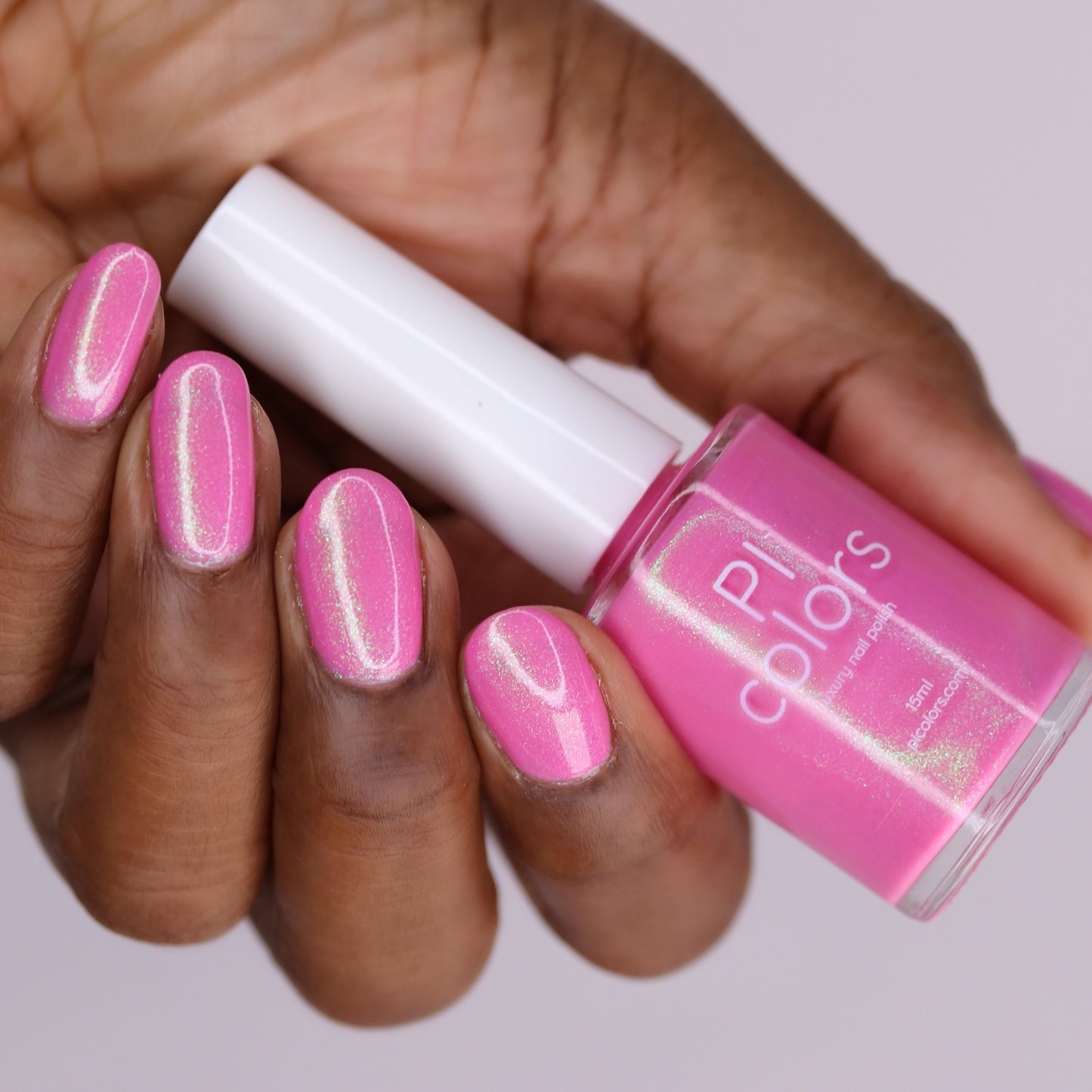 A hand with medium-dark skin holds a bottle of PI Colors' "Mint and Summer.100" handmade fluorescent pink nail polish, with nails painted in the same shimmery shade against a softly blurred light background.