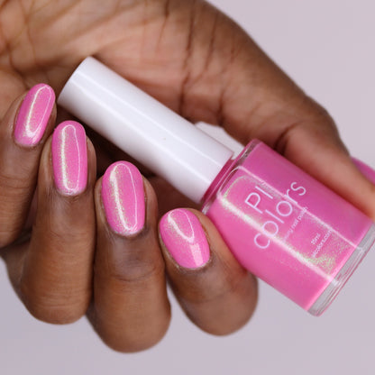 A hand with medium-dark skin holds a bottle of PI Colors' "Mint and Summer.100" handmade fluorescent pink nail polish, with nails painted in the same shimmery shade against a softly blurred light background.