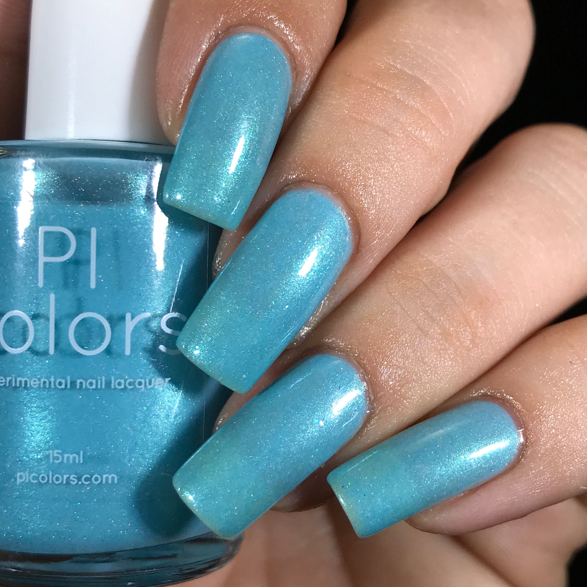 A close-up of a hand with long nails painted in a glossy shade of Berry Mousse.001 Blue Nail Polish, holding the bottle matching the polish. The bottle, complete with a white cap and labeled "PI Colors," adds an exquisite touch that enhances the mesmerizing effect.