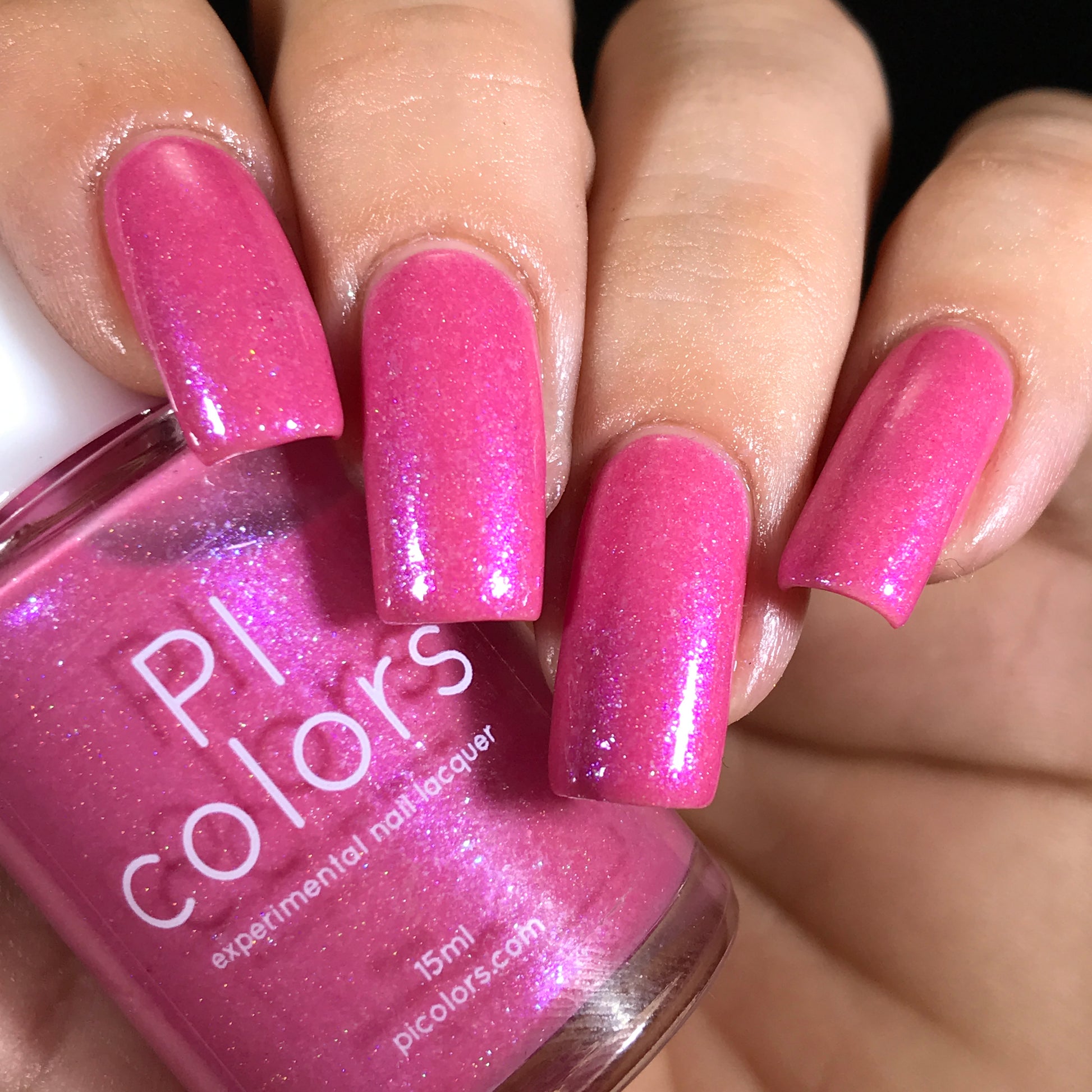 A hand with long, neatly manicured nails dazzles in shimmery pink, enhanced by holographic powder. The person holds a bottle of PI Colors' Strawberry Sundae.002 Pink Nail Polish, perfectly matching the glimmering shade on their nails.