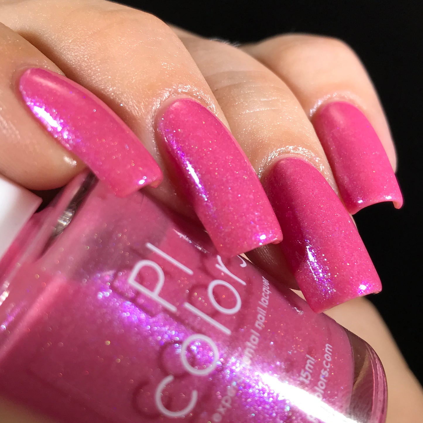 A close-up shot showcases a hand with long, shimmery pink nails adorned with holographic powder. The nail polish bottle labeled "Strawberry Sundae.002" by PI Colors perfectly complements the vibrant hue on the nails. The glossy finish stands out against a dark background, emphasizing its handmade quality.