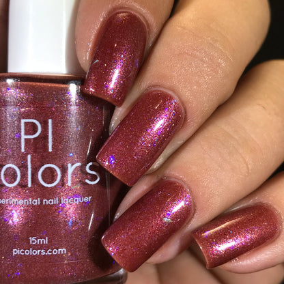 Close-up of a hand with long, square-shaped nails painted in Quasar.156 Red, a shimmery deep red shade. The luxury nail lacquer bottle is held in the hand and labeled "PI Colors" with a 15ml size indication.