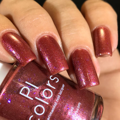 Close-up of a hand displaying nails adorned with Quasar.156, a shimmering metallic red nail polish that features color-shifting flakies. The luxury bottle of PI Colors nail lacquer is clasped between the thumb and index finger, perfectly matching the dazzling nail color.