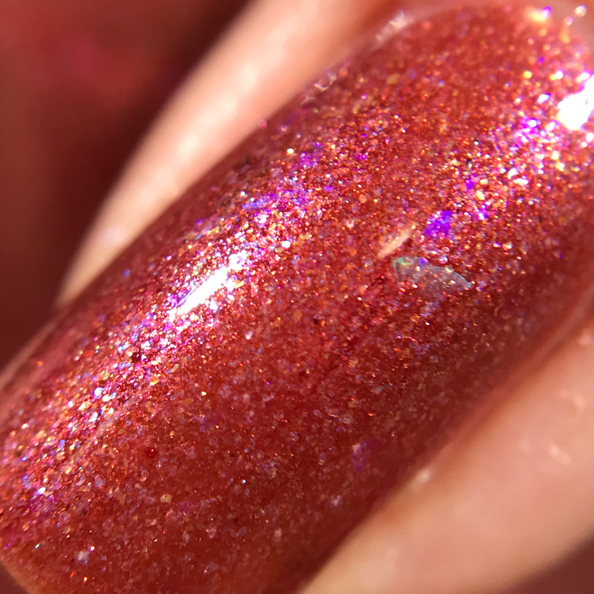 A close-up of a fingernail showcasing Quasar.156 Red Nail Polish by PI Colors in a luxurious glossy red-orange shade, highlighted with shimmering glitter and a subtle purple iridescence. The smooth texture is accentuated by color-shifting flakies for a reflective, sparkling finish.