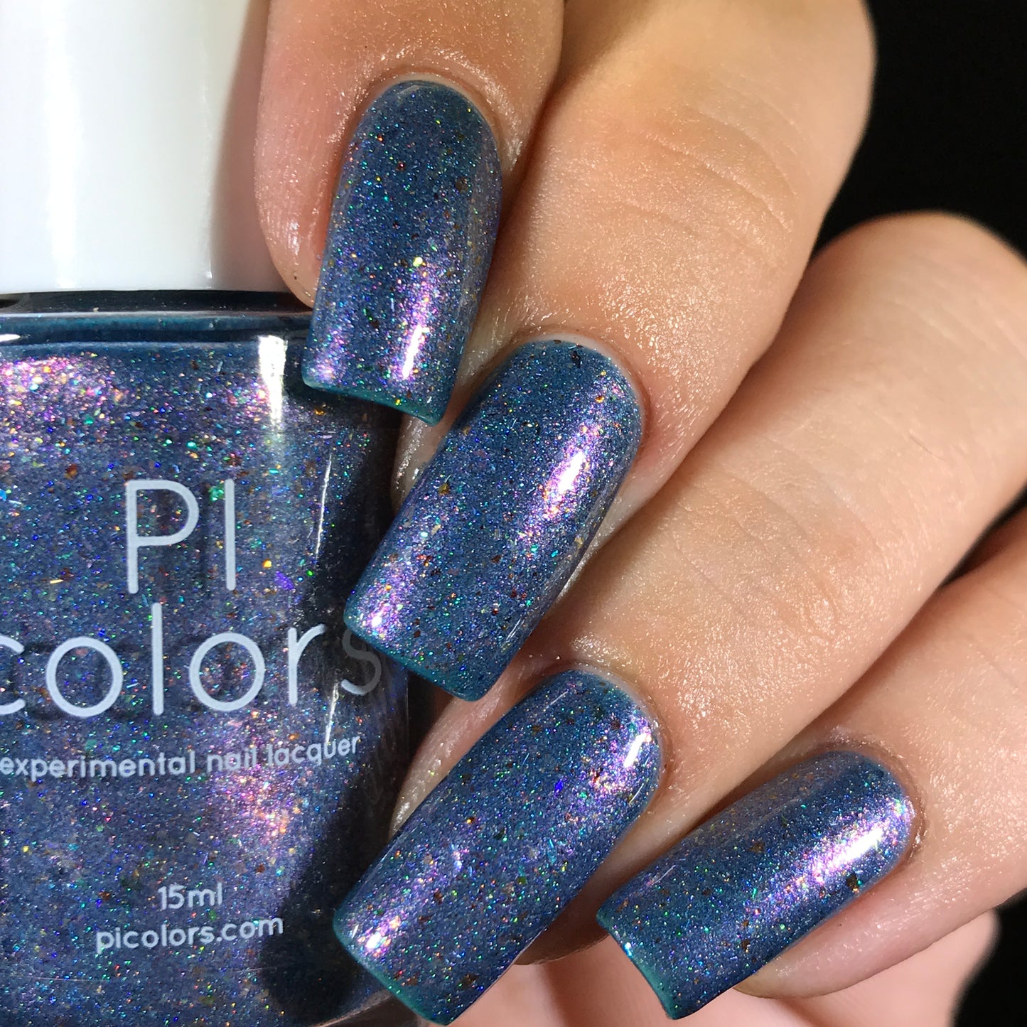 A close-up shows a hand with multicolored, glittery blue nails accentuated by deep blue polish, holding a bottle labeled "East of the Storm.029 Blue Nail Polish" by PI Colors. The experimental nail lacquer features color-shifting iridescent flakies and contains 15ml, available at picolors.com.