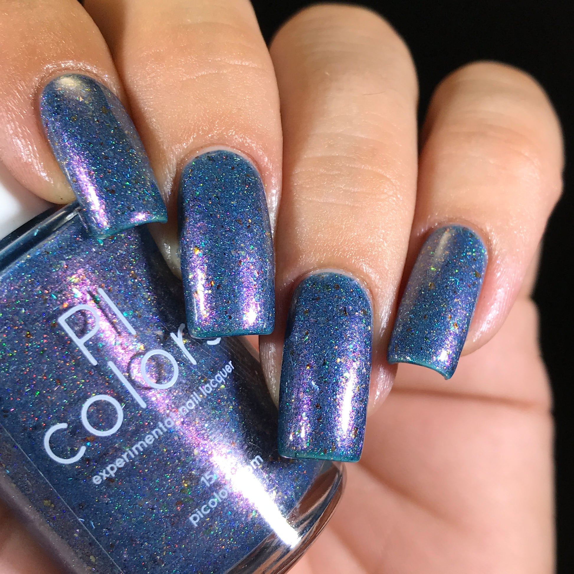 A hand features long fingernails coated with a deep blue polish embellished with shimmering iridescent flakes, reflecting purple and multicolored sparkles under the light. A bottle of East of the Storm.029 Blue Nail Polish by PI Colors is visible in the image, highlighting its captivating color-shifting effect.