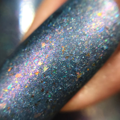 A close-up of the shimmering PI Colors East of the Storm.029 Blue Nail Polish on a fingernail highlights UCC flakies with blue, pink, and orange sparkles. The color-shifting iridescent finish creates a dazzling display as it catches light from different angles.