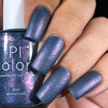 A hand with deep blue nails featuring color-shifting iridescent flakies holds a bottle of nail polish. The polish bottle has text partially visible, displaying "PI Colors" with the product name East of the Storm.029 Blue Nail Polish not fully readable.