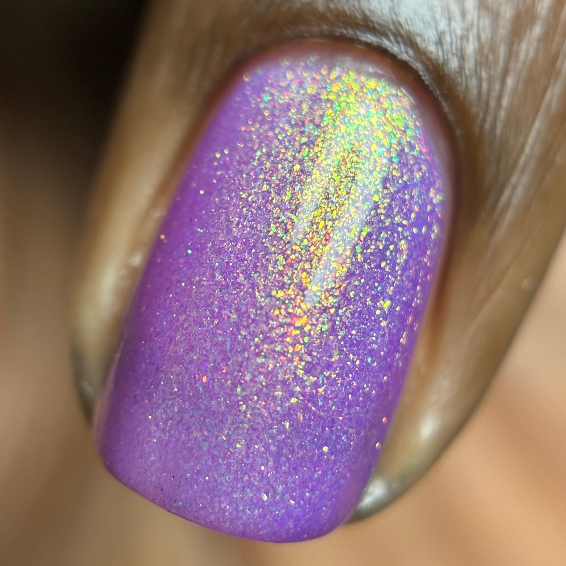 A close-up of a fingernail painted in a shimmery lavender-purple glitter polish with holographic sparkle reflecting gold, green, and pink tones.