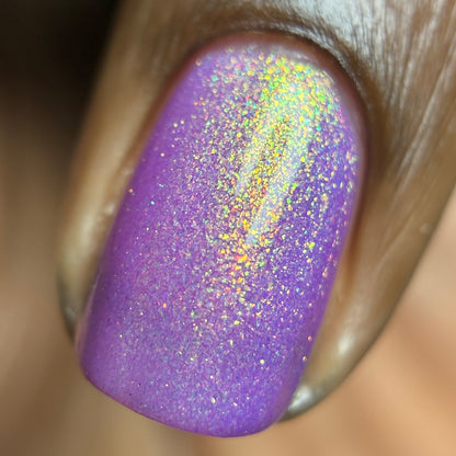 A close-up of a fingernail painted in a shimmery lavender-purple glitter polish with holographic sparkle reflecting gold, green, and pink tones.