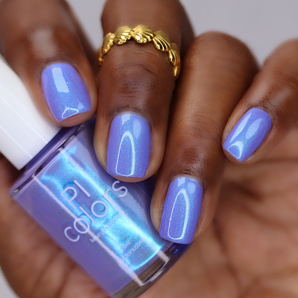 A close-up of a hand with glossy lavender-blue manicured nails holding a bottle of PL colors nail polish. The bottle label reads 'lavender blue shimmer' and the brand 'PL colors' is visible.