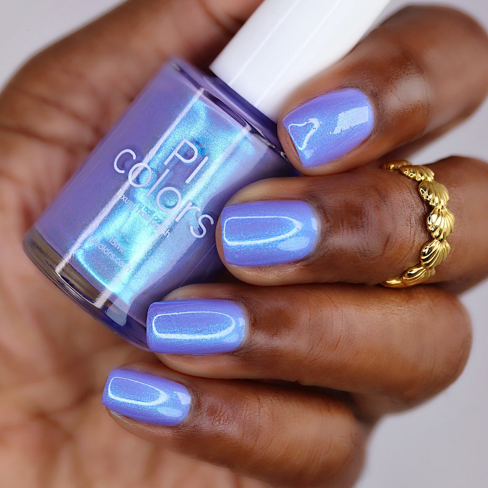 A close-up of a hand with lavender-blue manicured nails holding a bottle of nail polish. The nail polish in the bottle is a shimmering light blue with a lavender-purple undertone.