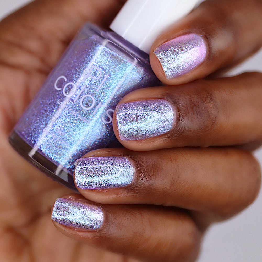 A close-up of a hand holding a bottle of nail polish. The nails are painted with a shimmering holographic polish that shifts between light purple, silver, and blue.