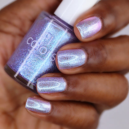 A close-up of a hand holding a bottle of nail polish. The nails are painted with a shimmering holographic polish that shifts between light purple, silver, and blue.