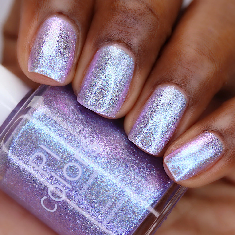 A close-up of a hand with manicured nails painted in a shimmering holographic lavender-pink nail polish, holding a bottle of the same polish.