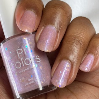 A hand with glossy, light pink Special Week.000 Nail Polish by PI Colors holds a similar bottle. This luxury lacquer features a subtle iridescent and glittery finish, adding a touch of sparkle to the nails.