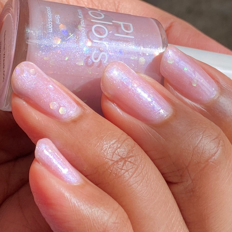 Close-up of a hand with light pink, shimmery nail polish from PI Colors' luxury line. This Special Week.000 Pink Nail Polish showcases subtle glitter and iridescent flakes, lending the nails a sparkling effect. The nails are neatly manicured, and the hand is holding the bottle of this exquisite polish.
