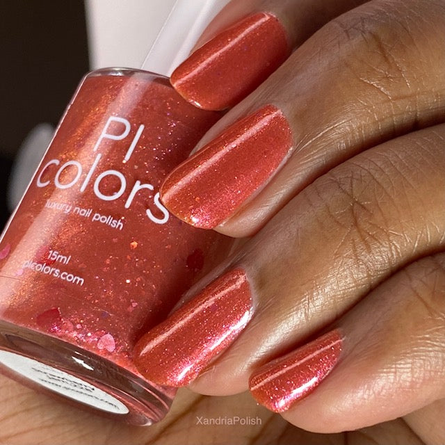 A close-up shot showcases a hand with manicured nails adorned in a shimmering coral shade. Between the fingers is a bottle of PI Colors' 10-free nail lacquer, labeled as "Strawberry Jam.228 Red Nail Polish," revealing its glittery coral hue. This handmade polish beautifully complements the warm brown skin tone.