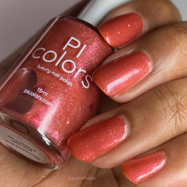 A close-up of a hand with neatly painted nails in a shimmering coral shade, holding a bottle of PI Colors luxury, handmade nail polish labeled "Strawberry Jam.228 Red Nail Polish," which matches the nail color and features the brand name and website.