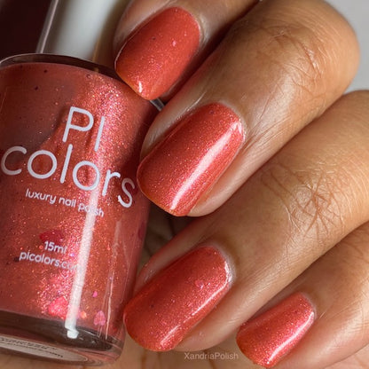 A hand with neatly manicured fingernails painted in a radiant crimson-red 10-free nail lacquer. The "PI Colors" bottle is held in the hand, harmonizing beautifully with the polished nails.