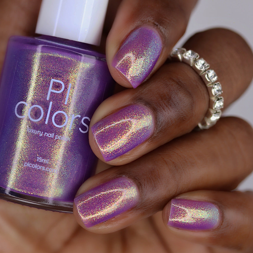 A close-up of a hand with nails painted in a shimmering holographic purple nail polish, holding a bottle of Picolors nail polish. The polish has an iridescent, shifting finish that reflects pink, gold, and silver tones.