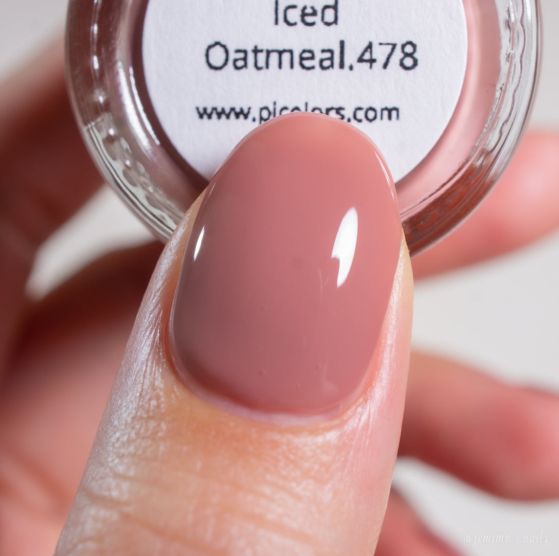 A close-up shows a fingernail painted with Iced Oatmeal.478, a glossy muted pink-nude shade by PI Colors. In the background, a jar labeled "Iced Oatmeal.478" from this 10-free brand is held, highlighting its official website.