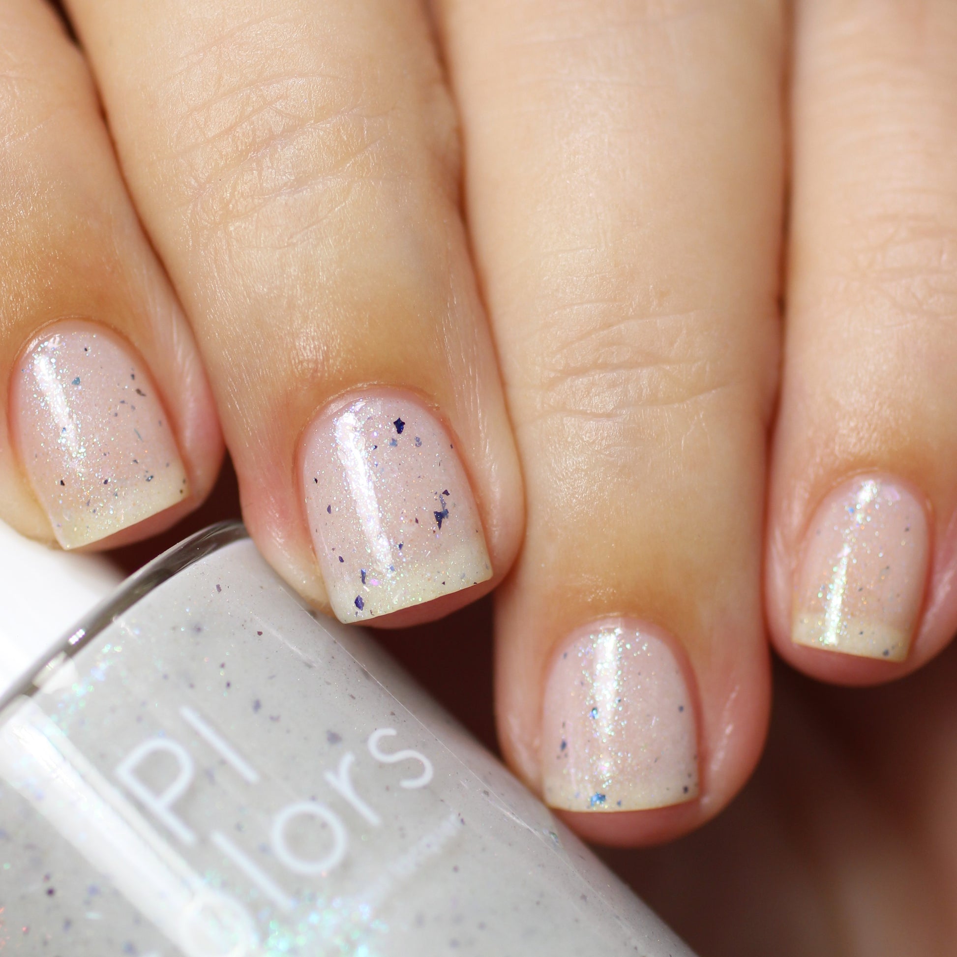 Close-up of manicured fingernails painted with PI Colors' Ionosphere.064 White Nail Polish, a glossy light pink hue that transforms with subtle blue and purple specks. A bottle of this handmade nail polish is partially visible in the foreground.