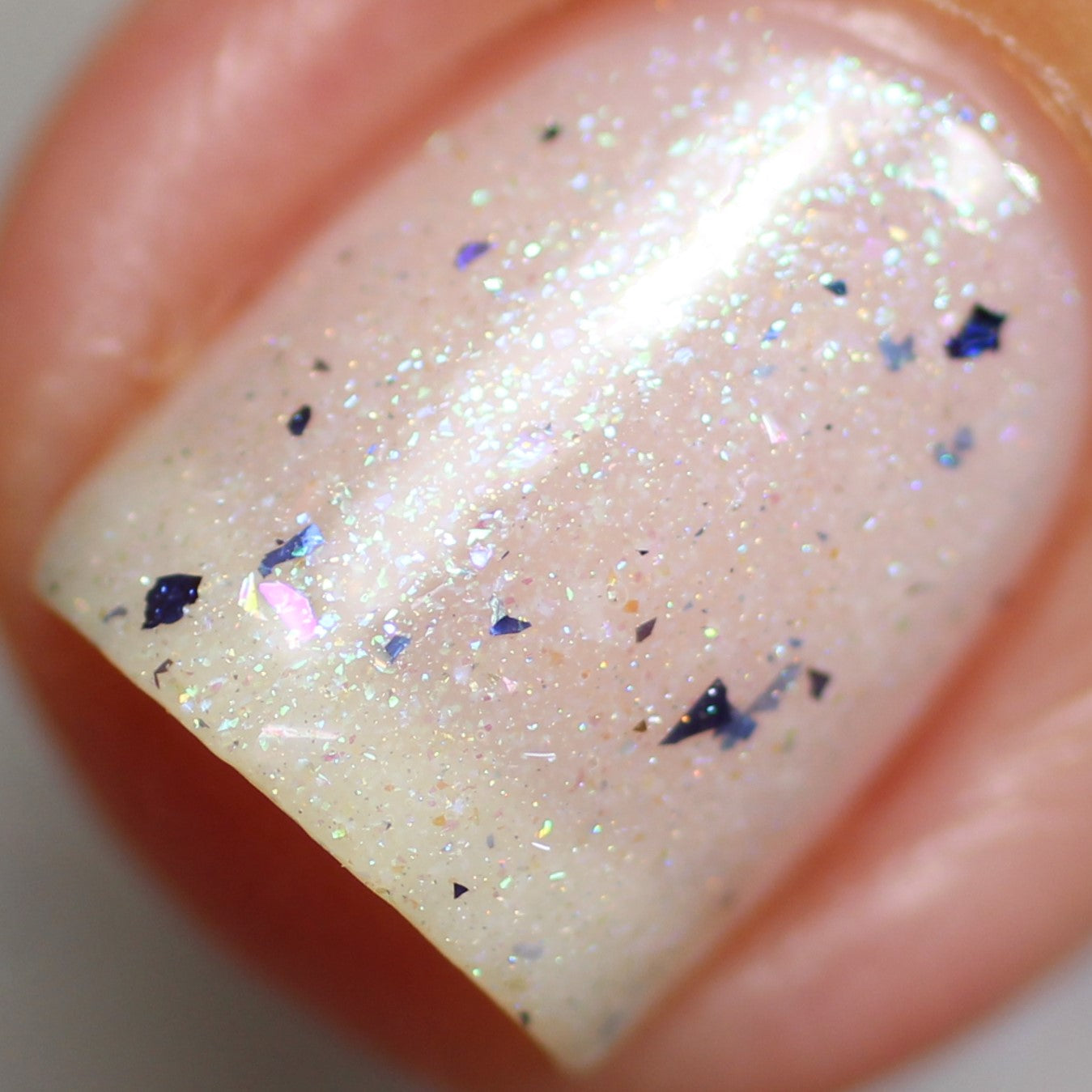 The photo features a close-up of a fingernail adorned with PI Colors' Ionosphere.064 White Nail Polish, a handcrafted shade in pale pink that boasts a shimmery, glittery finish with tiny multicolored flakes. The surface catches the light beautifully, showcasing its iridescent sparkle and subtle color-changing effect.