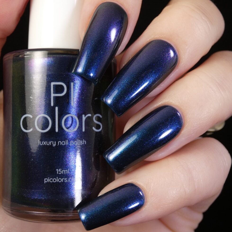 A hand with long nails painted in shimmering dark blue nail polish holds a bottle of PI Colors' luxury nail polish, Judge None Choose One.000. This handmade product boasts a matching dark blue hue with a metallic sheen, capturing 90s Nostalgia through its striking multichrome finish.