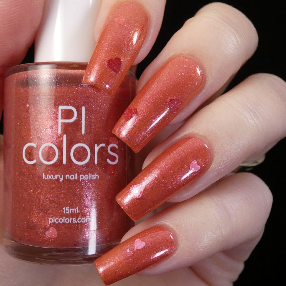 A hand with long nails painted in a shimmering red nail polish with heart-shaped glitters exudes elegance. Beside the nails, a bottle labeled "PI Colors" holds this luxurious "Strawberry Jam.228" nail lacquer.