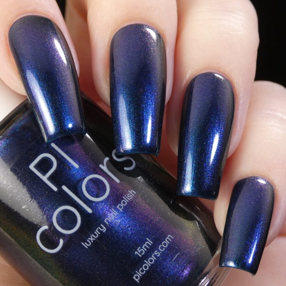 A close-up of a hand with long, manicured nails painted in a glossy, dark blue multichrome finish. A bottle of PI Colors' "Judge None Choose One.000" nail polish is held between the fingers, reflecting light and evoking 90s nostalgia.