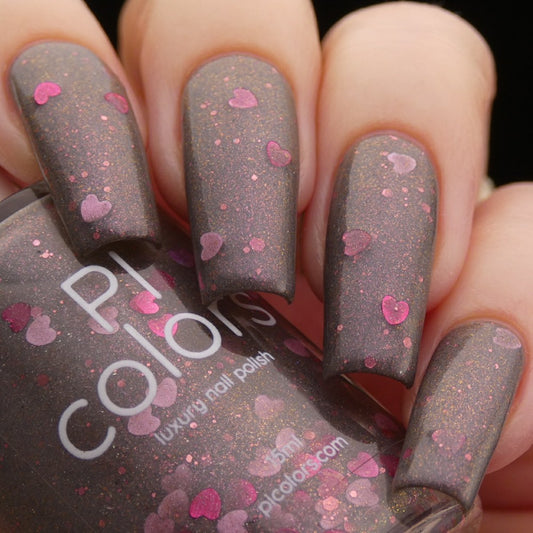A close-up shot highlights a hand with nails adorned in Chaos Heart.000 Gray Nail Polish, a dark brown handmade polish embellished with pink heart-shaped glitter. The partially visible bottle displays "PI Colors" in white text, and the shimmery finish lends it a luxurious appearance.
