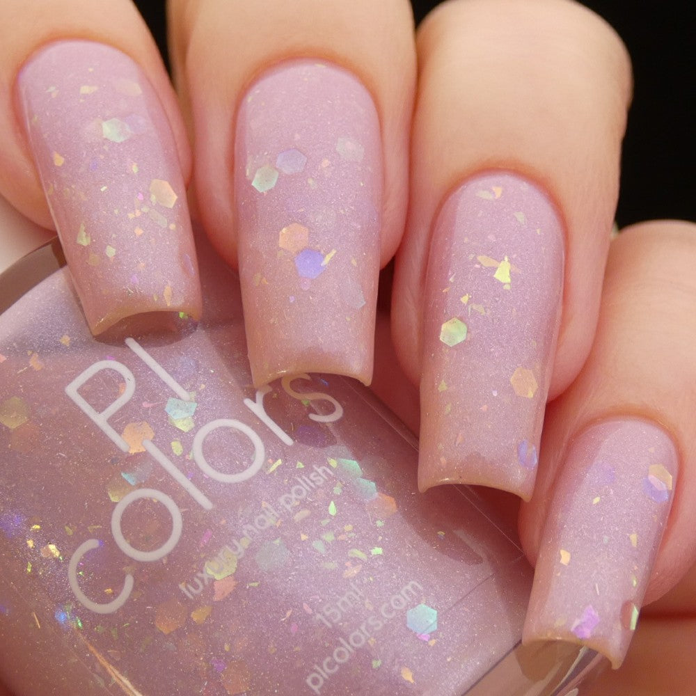 Close-up of a hand with long nails adorned in Special Week.000 Pink Nail Polish, a pastel pink hue filled with iridescent rainbow flakies. In the hand is the matching bottle of this handmade nail polish, proudly showcasing the PI Colors brand. The whimsical design captures the charm and flair reminiscent of Uma Musume Pretty Derby.