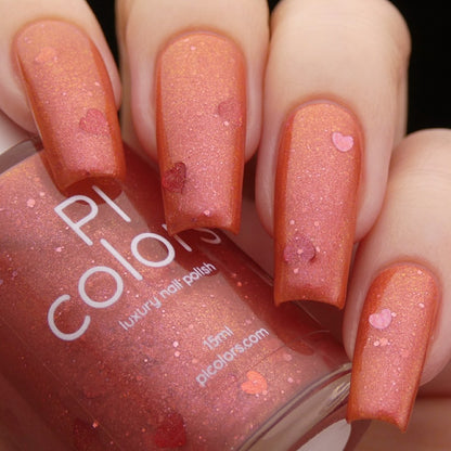 Close-up of a hand with long nails painted in a shimmery orange nail polish adorned with small, heart-shaped red glitter. The hand holds a matching bottle of "Strawberry Jam.228 Red Nail Polish" from the luxury brand PI Colors.