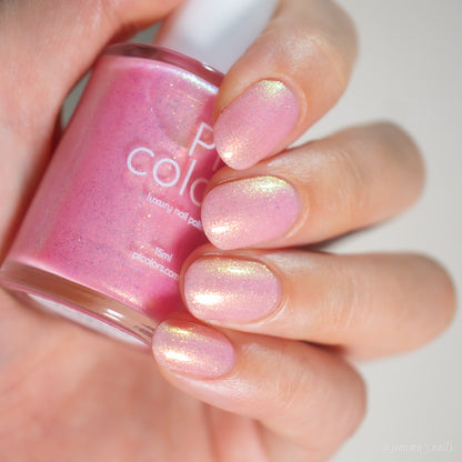 A hand with short nails painted in Lemon Pink Sugar Cookie.422 by PI Colors holds a matching handmade bottle of this luxury light pink polish, featuring an iridescent golden shimmer and the visible brand name.