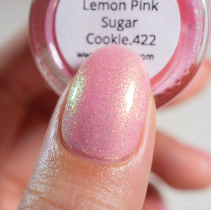 A close-up of a thumb nail painted with PI Colors' Lemon Pink Sugar Cookie.422, a shimmery handmade light pink polish featuring fine glitter and an iridescent sheen. The labeled jar is visible in the background.