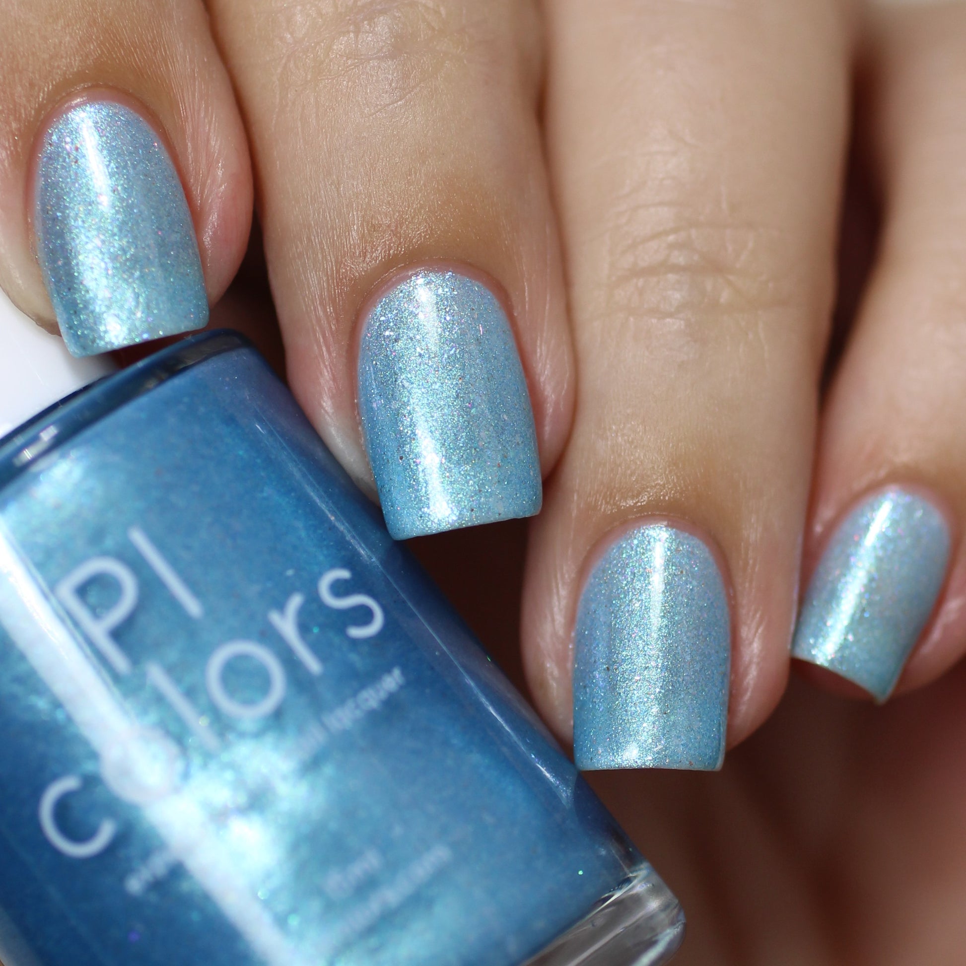 A close-up shot captures hands with neatly manicured nails adorned in Loulaki.101 Blue Nail Polish by PI Colors, showcasing iridescent flakies. The bottle of this handmade nail polish is held in the image, providing the nails with a glossy, sparkling finish.