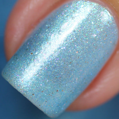 Close-up of a fingernail painted with PI Colors' Loulaki.101 Blue Nail Polish, a handmade shimmery blue polish highlighted by iridescent flakies that provide a fine glittery texture. The polish captures light beautifully, resulting in a sparkling effect against the blurred background.