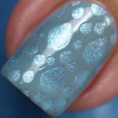 Close-up of a fingernail adorned with shimmering light blue nail polish from PI Colors' Loulaki.101 collection. This handmade polish is enriched with iridescent flakies that create reflective, glittery droplets resembling a raindrop pattern on the light blue backdrop. The finish appears both glossy and smooth.