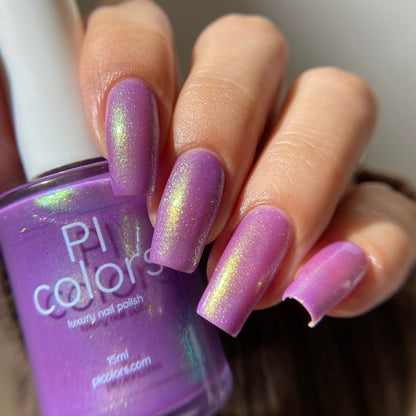 A close-up of a hand with nails painted in a lavender purple polish with an iridescent shimmer, holding a bottle of P! colors luxury nail polish.