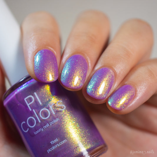 A close-up of a hand with nails painted in an iridescent purple nail polish with gold shimmer, holding a bottle of PI colors luxury nail polish.