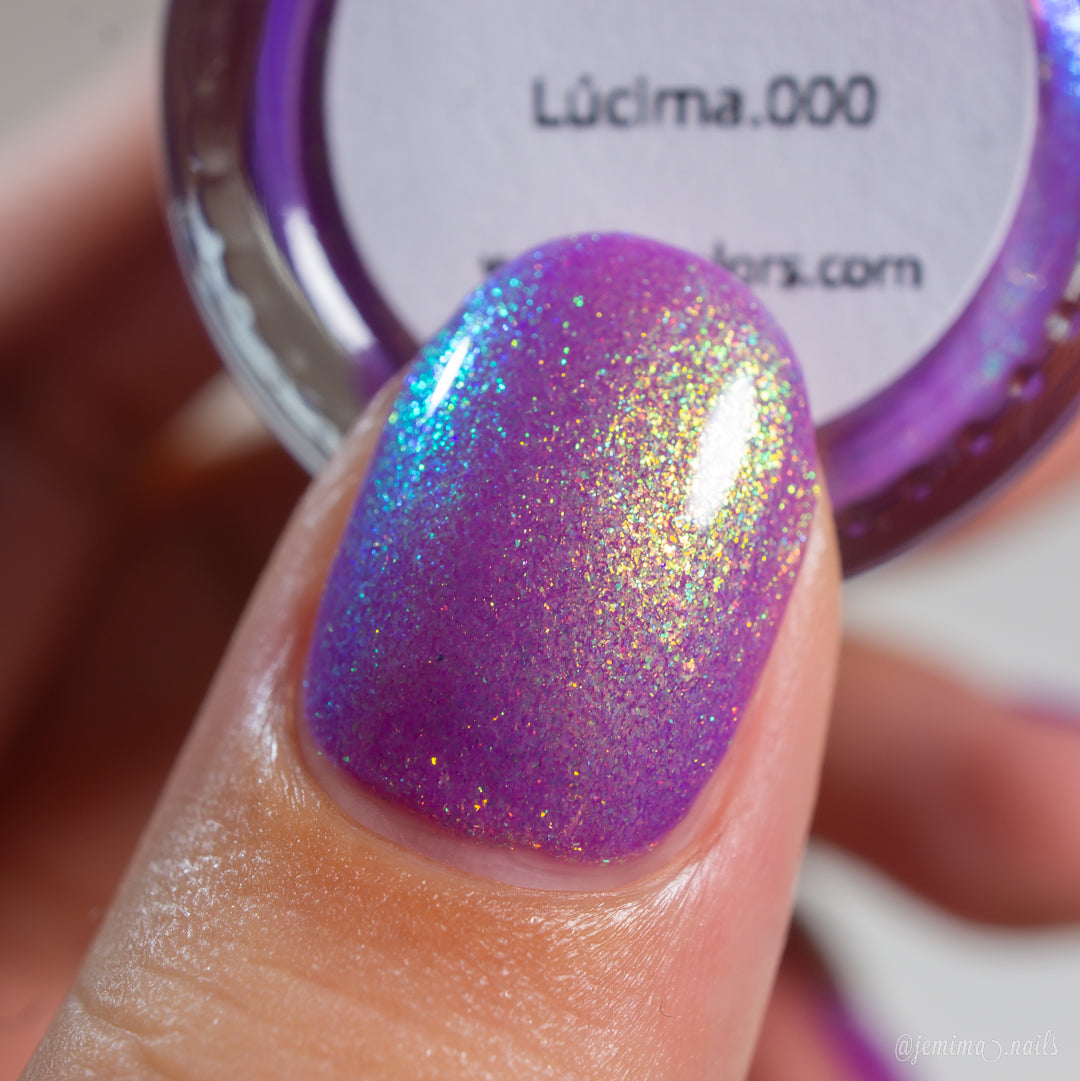 A close-up of a fingernail painted with a sparkling holographic purple nail polish, with a small open jar of the same polish in the background. The label reads 'Lüi.