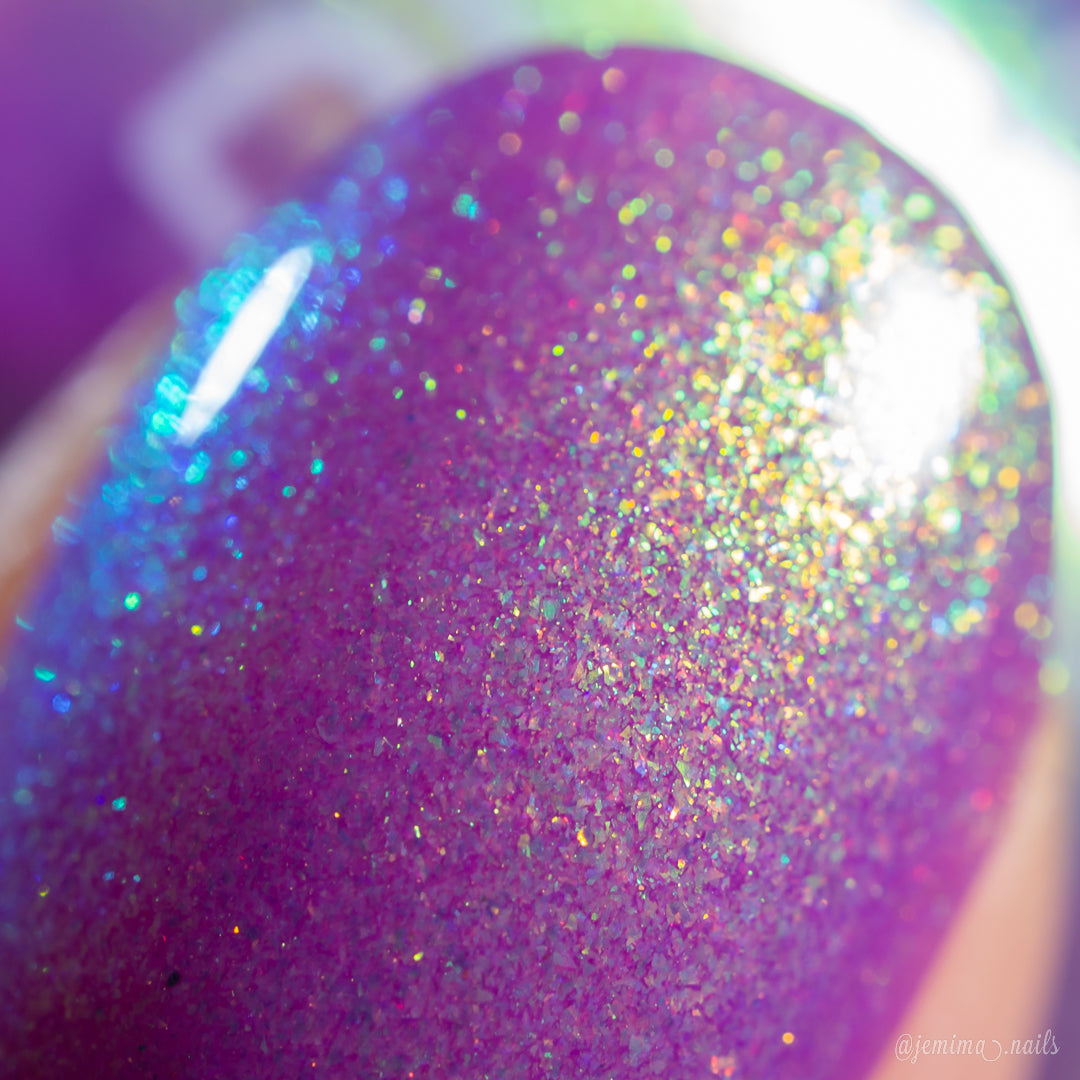A close-up of a nail tip painted with a glittery purple nail polish that has strong holographic iridescent shimmer, reflecting blue, green, and gold tones.