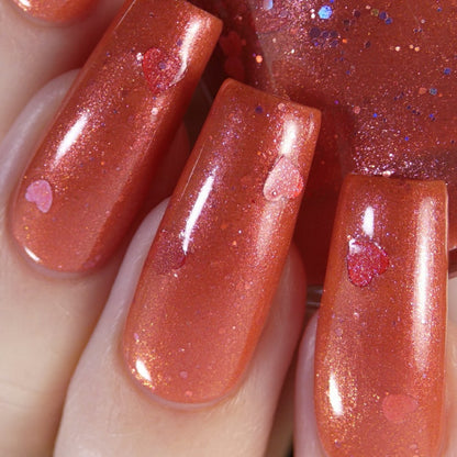 Close-up of fingernails painted in Strawberry Jam.228 Red Nail Polish by PI Colors, featuring a luxurious, shimmering orange-pink hue with tiny heart-shaped flecks. The nails are neatly manicured, and the handmade nail polish provides a glimmering effect under the light.